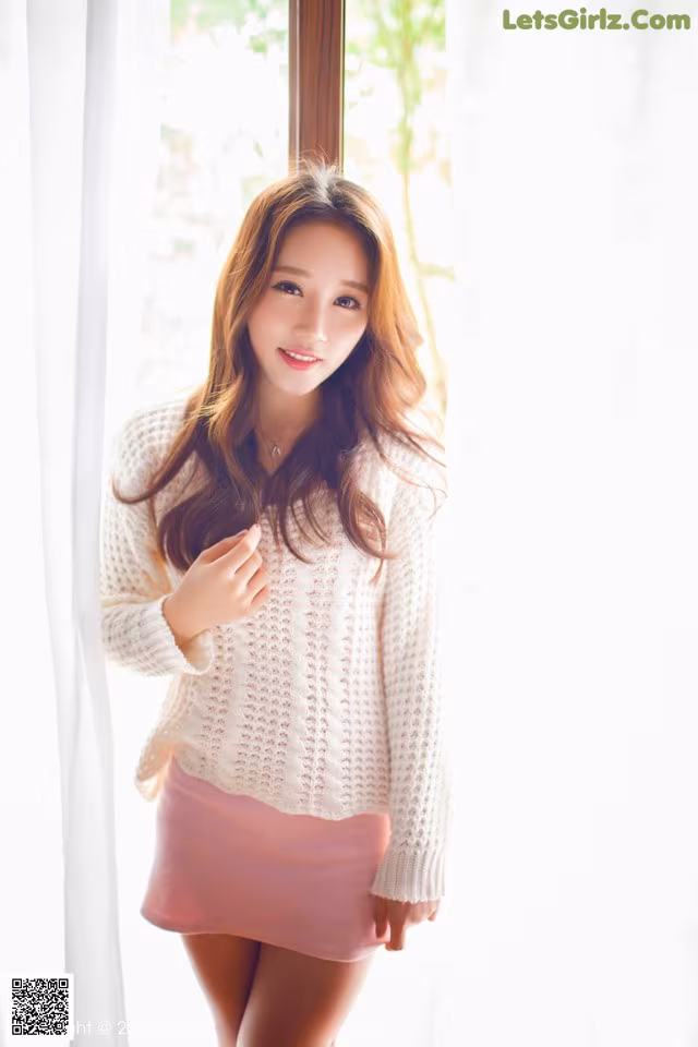 A woman in a white sweater and pink skirt posing by a window.