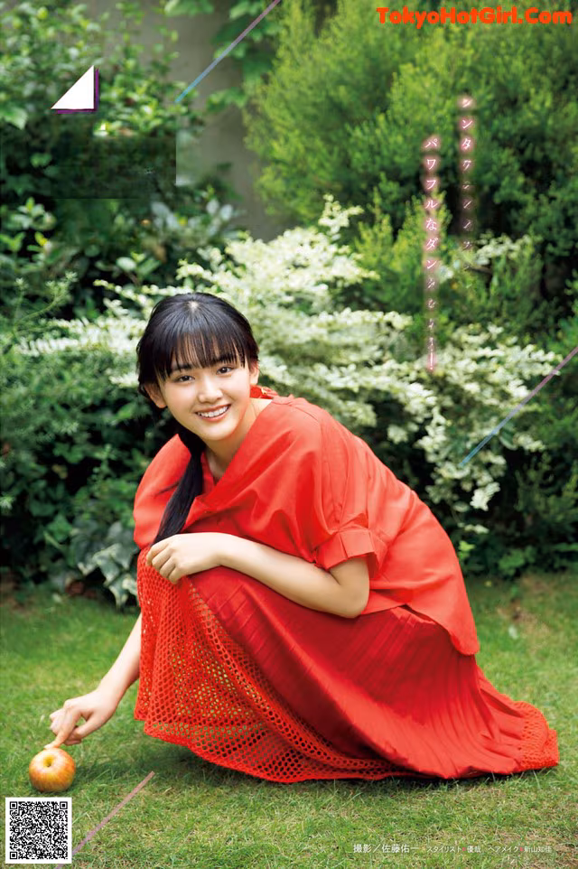 A woman in a red dress sitting on the grass with an apple.