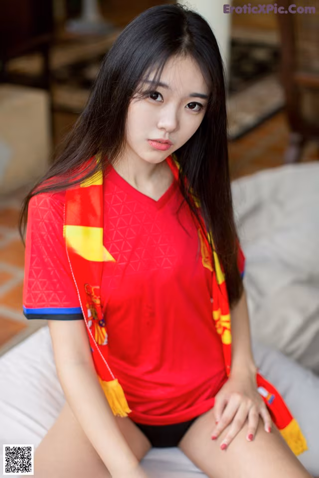 A woman in a red shirt sitting on a bed.